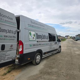 DomyJutra.pl Piotr Tereszkiewicz - Szary samochód dostawczy z otwartymi drzwiami, oklejony reklamą firmy budującej domy energooszczędne i pasywne, na tle placu budowy i rzędu nowych domów.