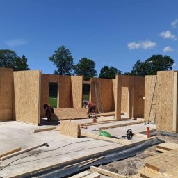 DomyJutra.pl Piotr Tereszkiewicz - Wznoszenie ścian domu szkieletowego z płyt OSB, widoczni pracownicy podczas montażu i cięcia elementów, na ścianie widoczne logo firmy Solcraft, w tle zieleń drzew i błękitne niebo.