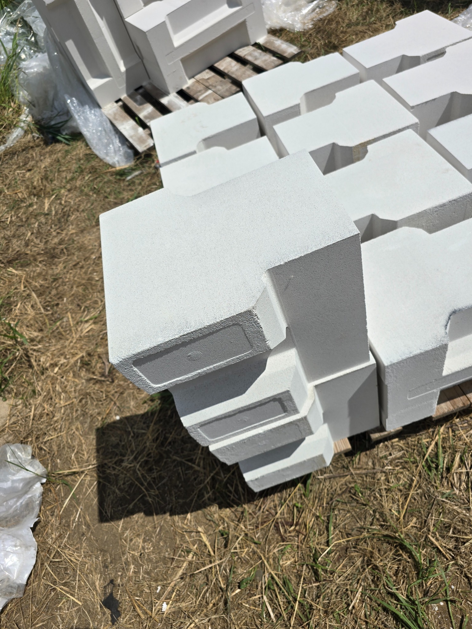 Paleta z białymi bloczkami fundamentowymi z wycięciami, ułożona na trawie i drewnianej palecie.