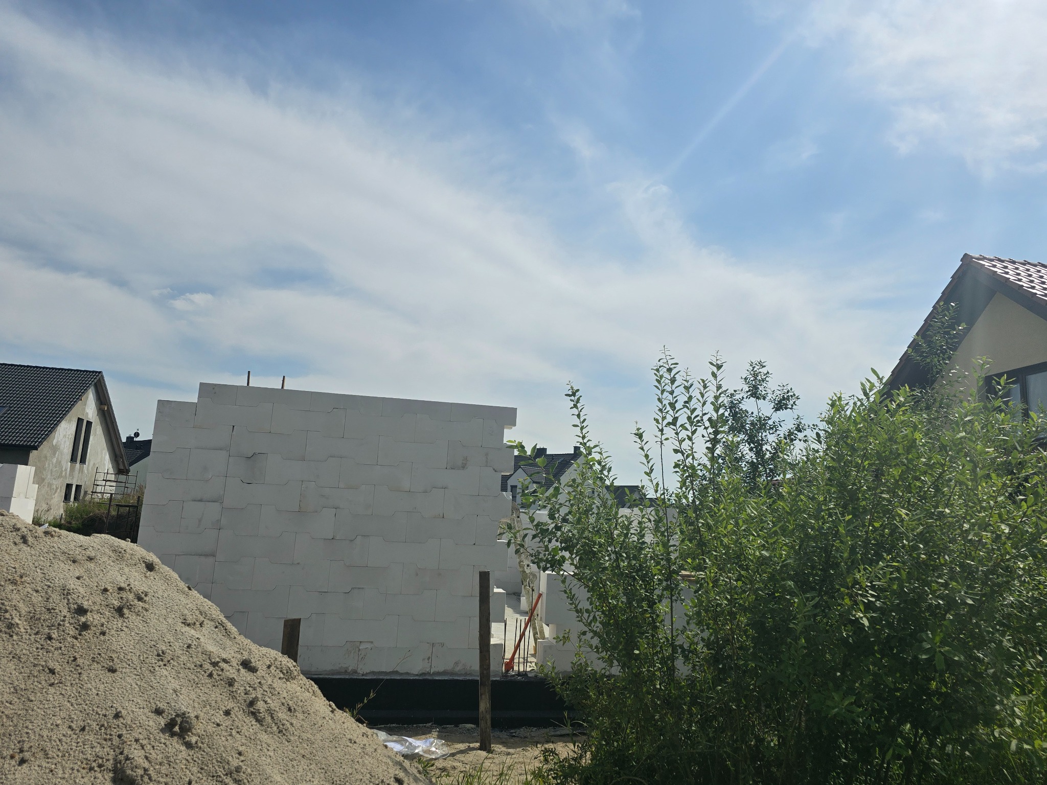 Wznoszone ściany z białych bloczków betonu komórkowego na budowie domu, widoczna sterta piasku i zieleń krzewów na pierwszym planie, błękitne niebo w tle.
