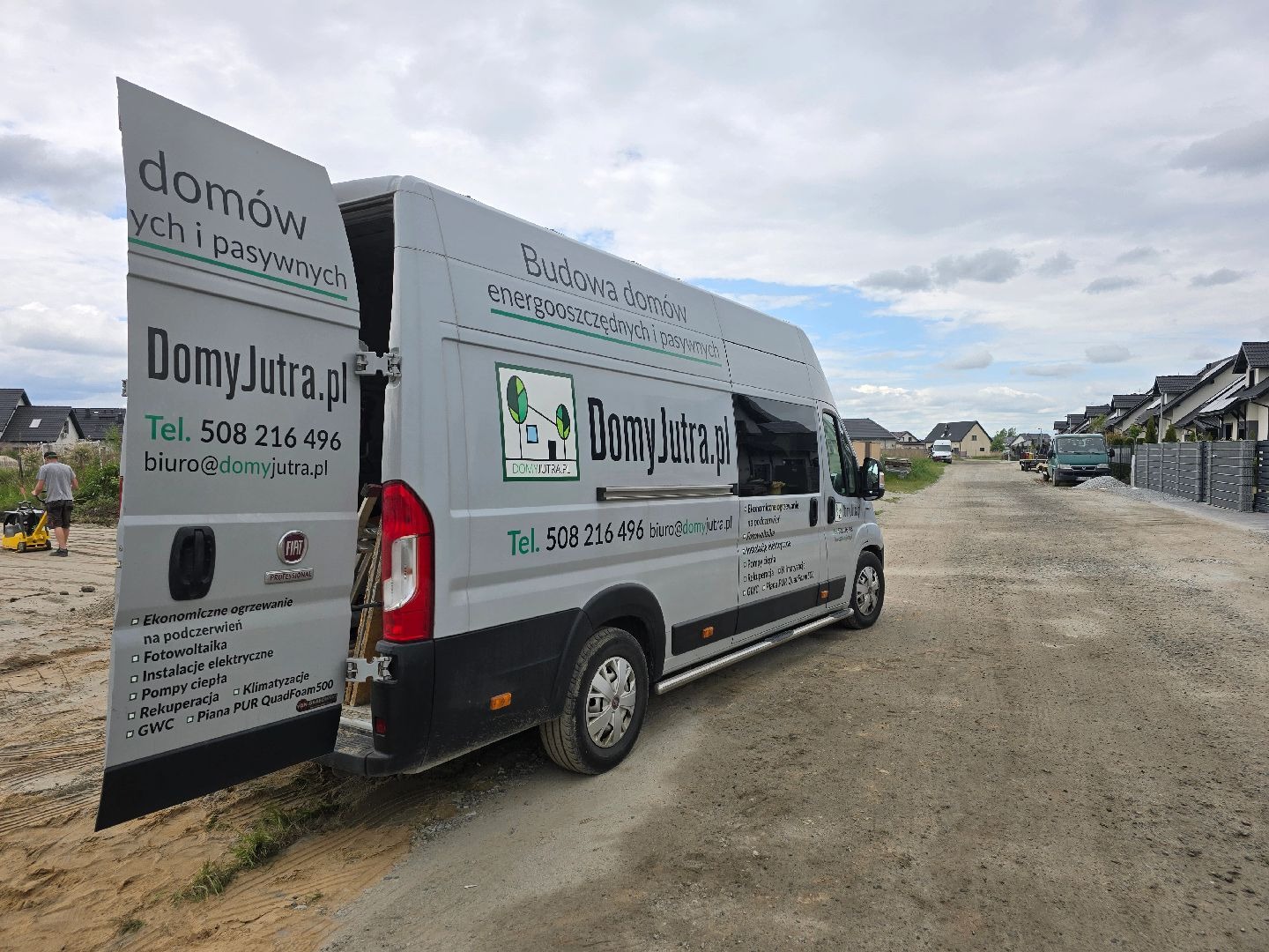 Szary samochód dostawczy z otwartymi drzwiami, oklejony reklamą firmy budującej domy energooszczędne i pasywne, na tle placu budowy i rzędu nowych domów.