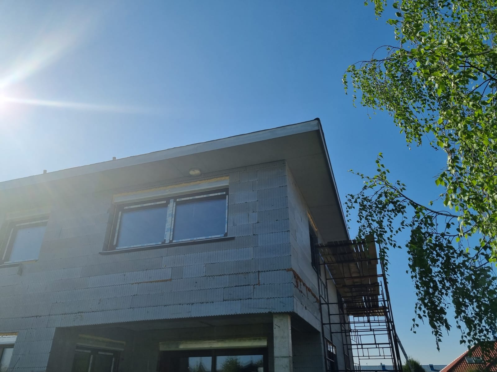 Surowy stan dwukondygnacyjnego budynku z szarej cegły, widoczne okna z folią ochronną i rusztowanie z boku, błękitne niebo i gałąź drzewa w tle.