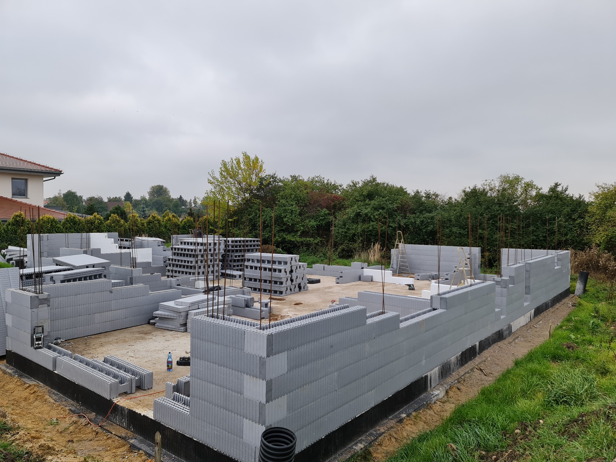 Budowa fundamentów z szarych bloczków betonowych na działce, widoczne wystające pręty zbrojeniowe, w tle drzewa i fragment domu sąsiada.