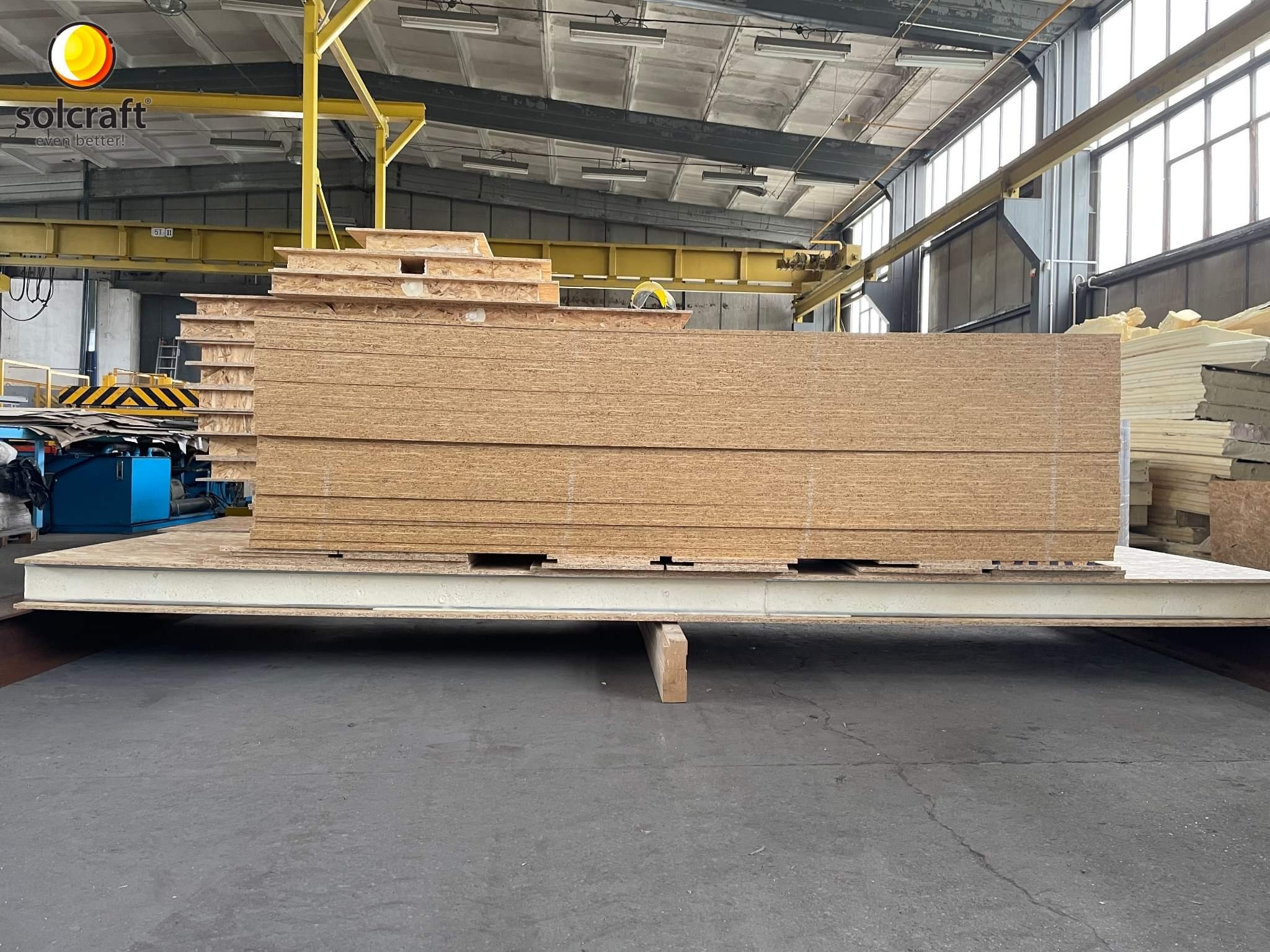 Duży stos płyt OSB na metalowej platformie w hali produkcyjnej, widoczny suwnica i logo producenta Solcraft.