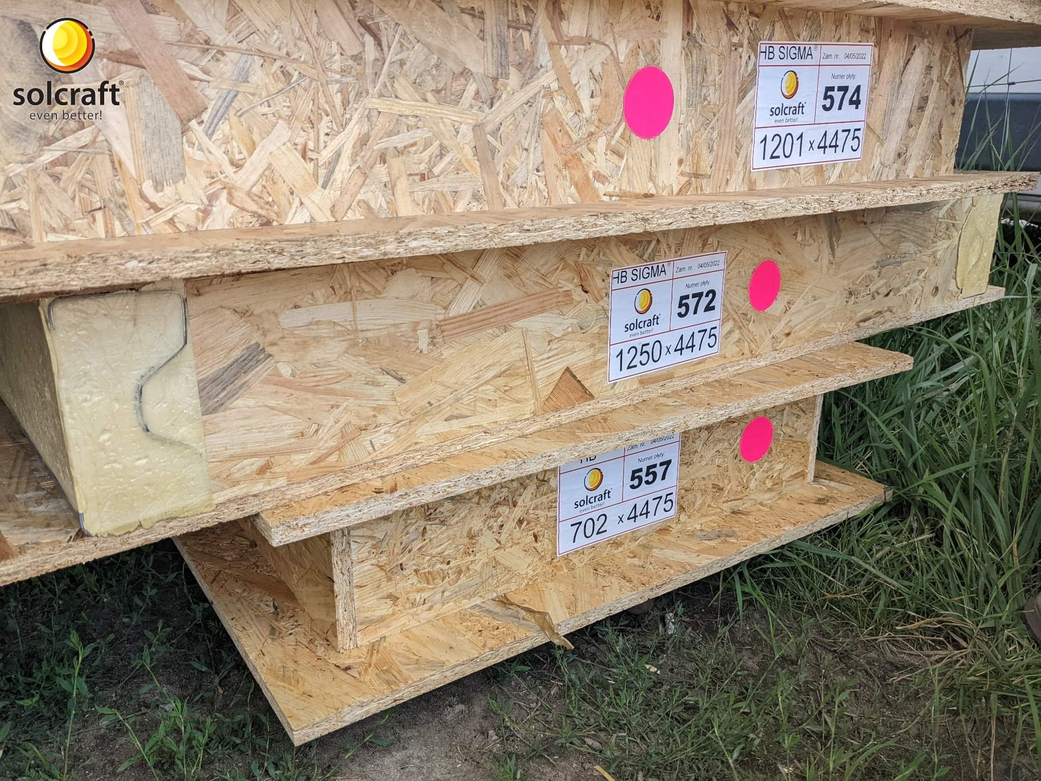 Płyty OSB w drewnianej konstrukcji z widocznymi etykietami producenta Solcraft i numerami płyt, zabezpieczone różowymi naklejkami, składowane na zewnątrz.