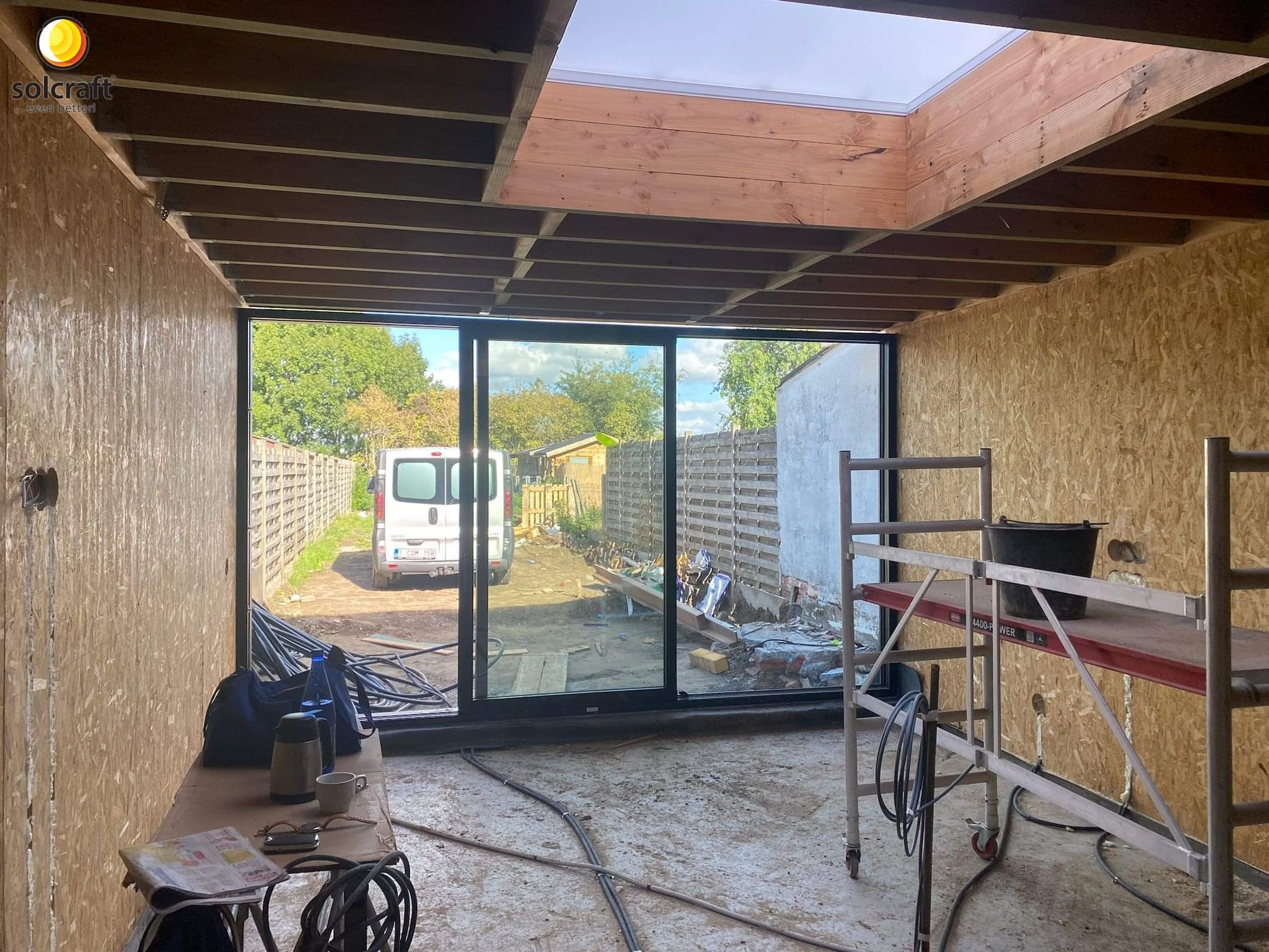 Wnętrze w trakcie budowy z drewnianą konstrukcją szkieletową, widoczne duże okno przesuwne z widokiem na ogród, rusztowanie i materiały budowlane wewnątrz.