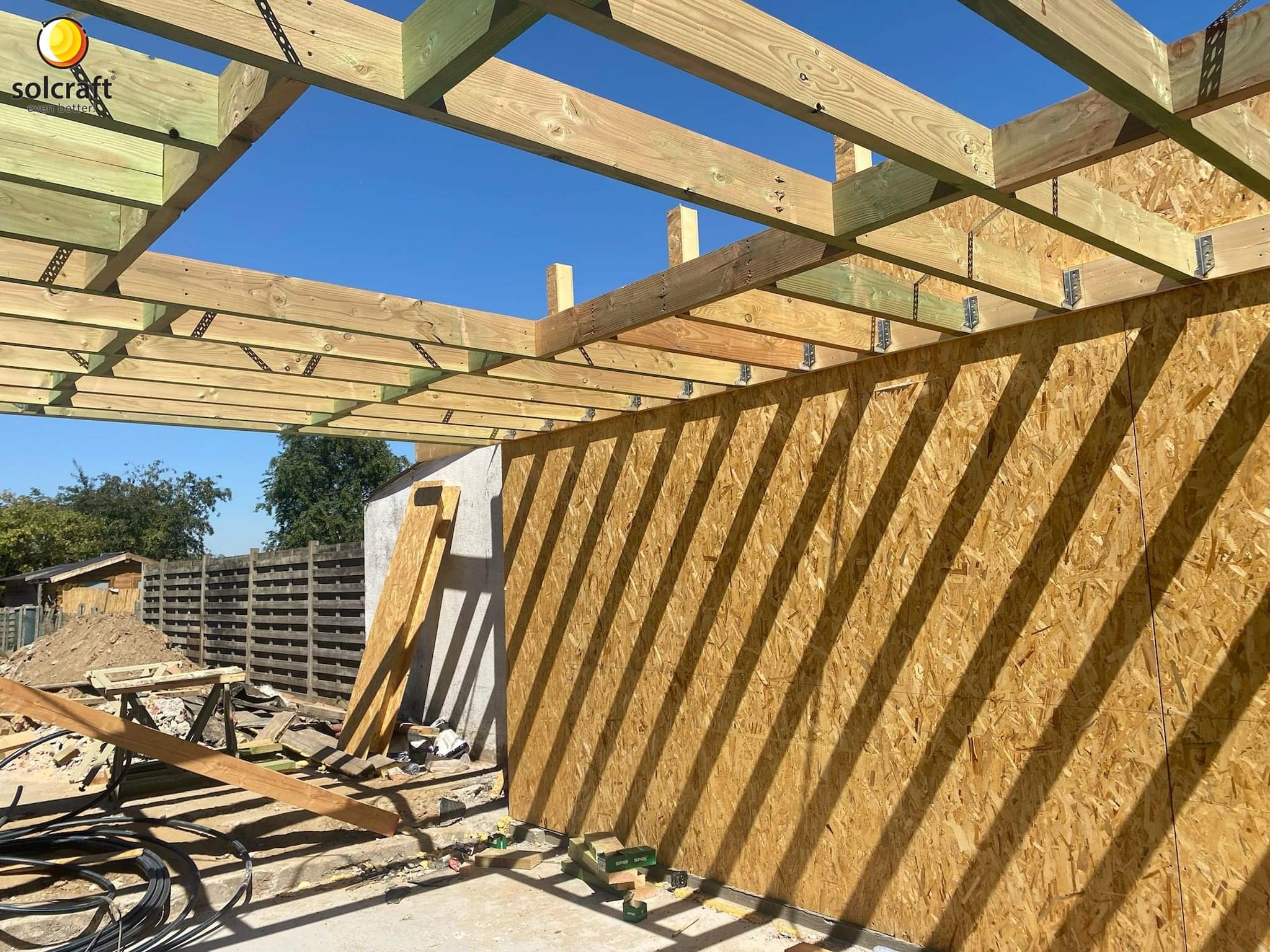 Wznoszenie konstrukcji budynku: drewniany szkielet dachu z widocznymi łącznikami stalowymi i poszycie ścian z płyt OSB w świetle słonecznym.