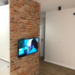 EMERWA Usługi wykończeniowe - Telewizor zawieszony na ścianie z cegły, obok zabudowy meblowej w jasnym pomieszczeniu z drewnianą podłogą i oświetleniem punktowym.