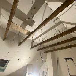 EMERWA Usługi wykończeniowe - Skośny sufit poddasza z drewnianymi belkami i zamontowanymi płytami gipsowo-kartonowymi, widoczne otwory na oświetlenie i okno dachowe, częściowo poprowadzone przewody elektryczne.
