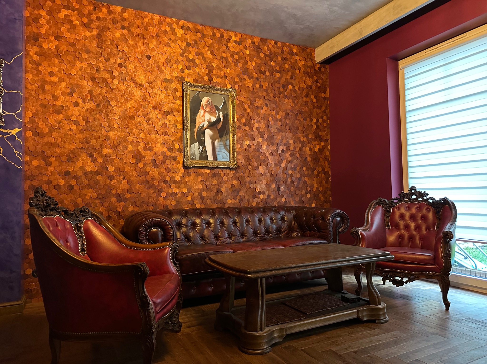 Elegancki salon z ciemnoczerwonymi ścianami, obitymi heksagonalnymi panelami, skórzanym chesterfieldem, dwoma fotelami w stylu Ludwika i obrazem w złotej ramie.