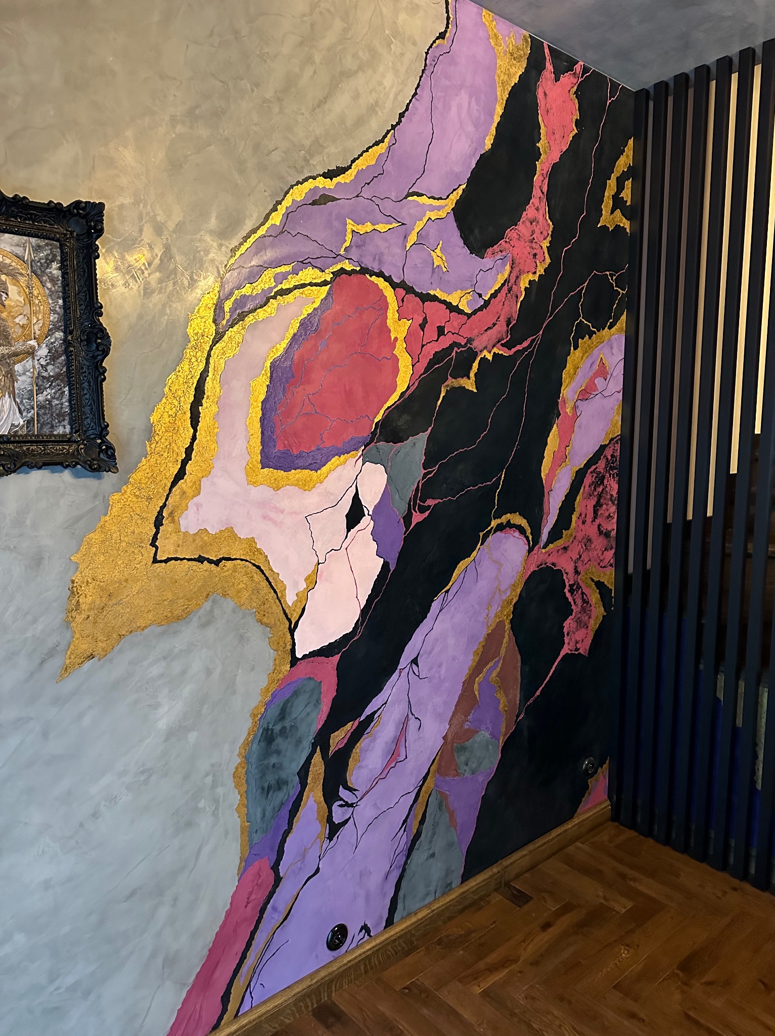 Dekoracyjna ściana z efektem marmuru w odcieniach różu, fioletu, czerni i złota, kontrastująca z szarą, fakturowaną powierzchnią i drewnianą podłogą w jodełkę, obok czarne listwy dekoracyjne.