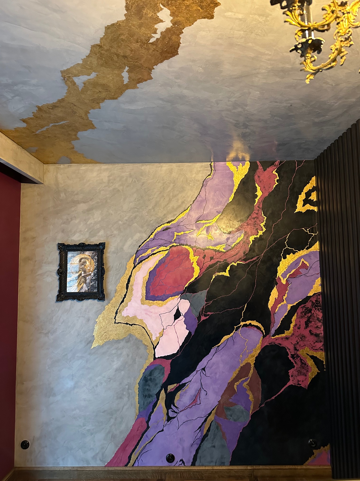 Dekoracyjna ściana z efektem marmuru w odcieniach czerni, fioletu i złota, połączona z szarą ścianą i złotymi akcentami na suficie, uzupełniona obrazem w ozdobnej ramie i nowoczesnym oświetleniem.