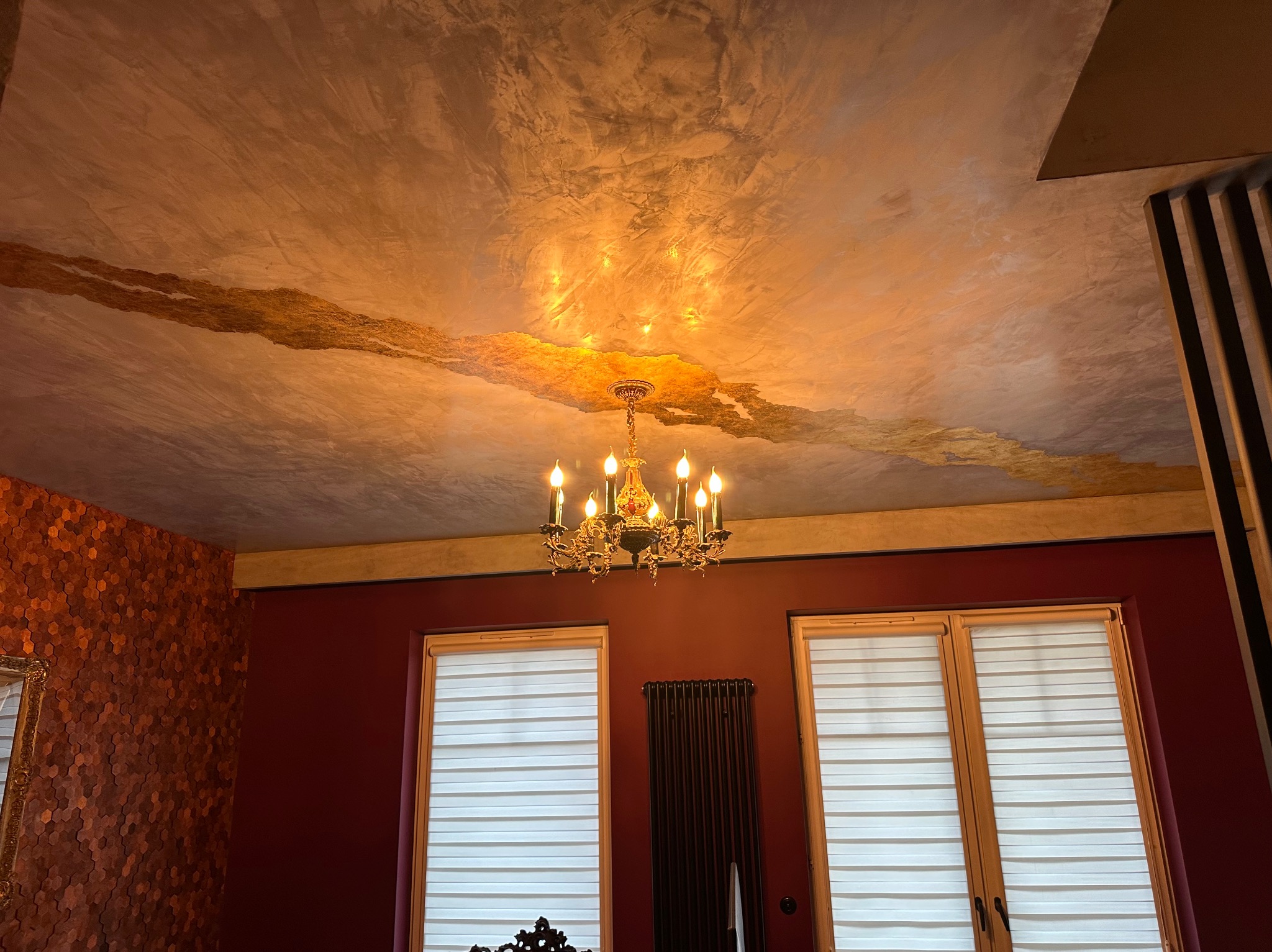 Dekoracyjny sufit z efektem marmuru i złotymi akcentami, oświetlony ozdobnym żyrandolem, widok na czerwone ściany z drewnianymi panelami i okna z roletami.