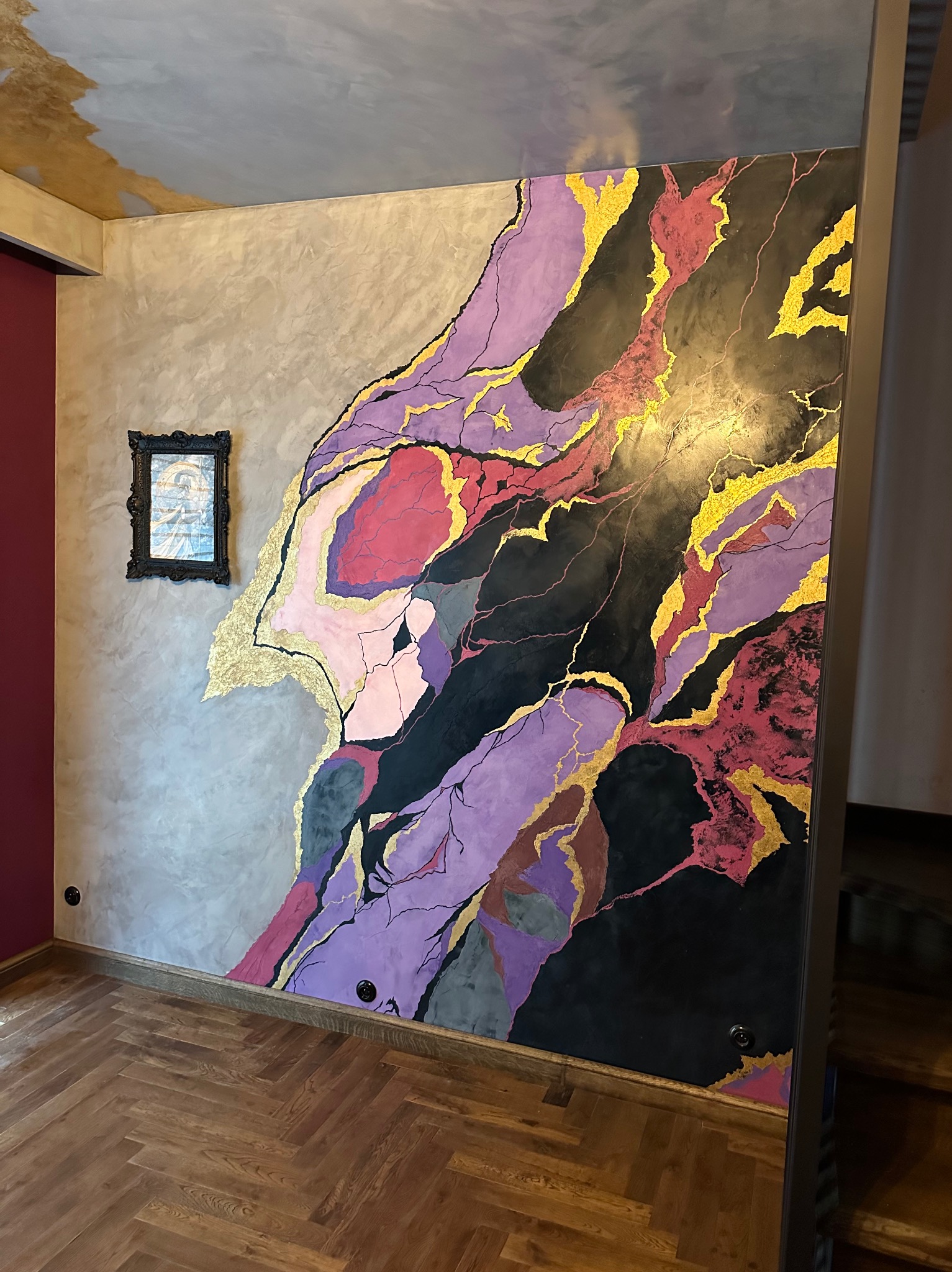 Dekoracyjna ściana z efektem marmuru w odcieniach szarości, fioletu, czerni i złota, ozdobiona ramką z obrazem, na podłodze parkiet ułożony w jodełkę.