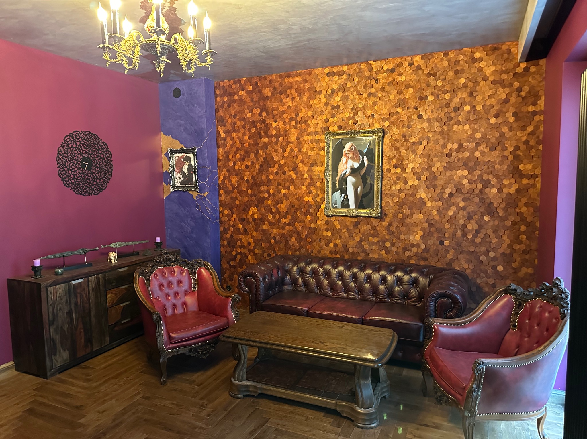 Salon w stylu eklektycznym z bordową sofą Chesterfield, dwoma czerwonymi fotelami Ludwik, drewnianą szafką RTV z dekoracjami, ścianą z heksagonalnych płytek i złotym żyrandolem.