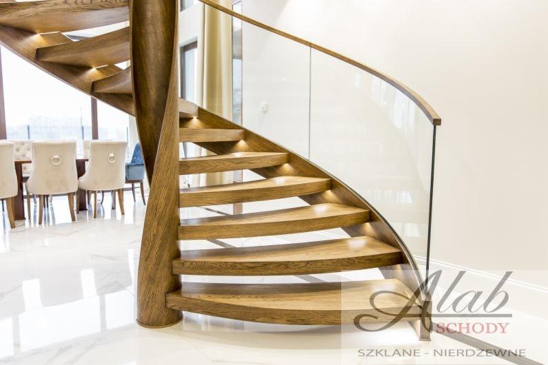 Spiralne schody z drewna z podświetleniem LED na stopniach i szklaną balustradą, widok na jadalnię w tle.