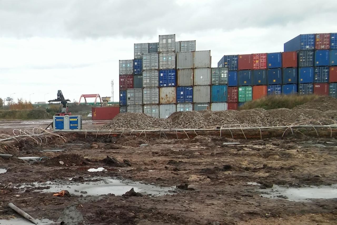 Teren budowy terminalu kontenerowego z widoczną maszyną do stabilizacji gruntu, rurami grzewczymi i stertą kontenerów w tle, pochmurne niebo.