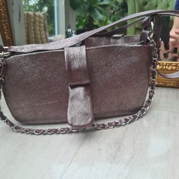 Budrem - Błyszcząca, bordowa torebka damska z łańcuszkiem, leżąca na podłodze z paneli, w tle widoczne rośliny i fragment lustra.