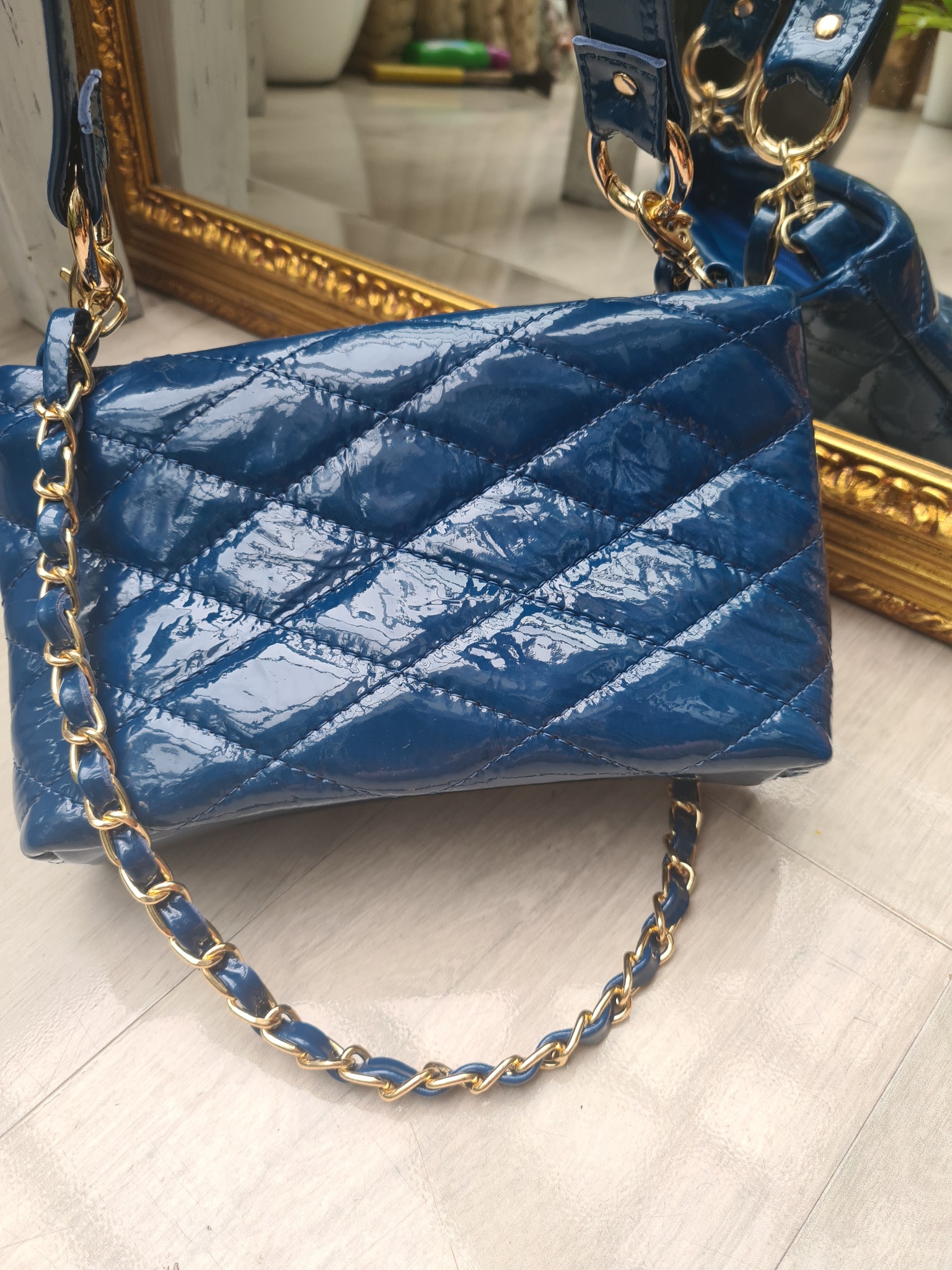 Błyszcząca, pikowana torebka w odcieniu głębokiego błękitu z złotym łańcuszkiem, ustawiona na jasnej podłodze z widocznym odbiciem w ozdobnym, złotym lustrze.