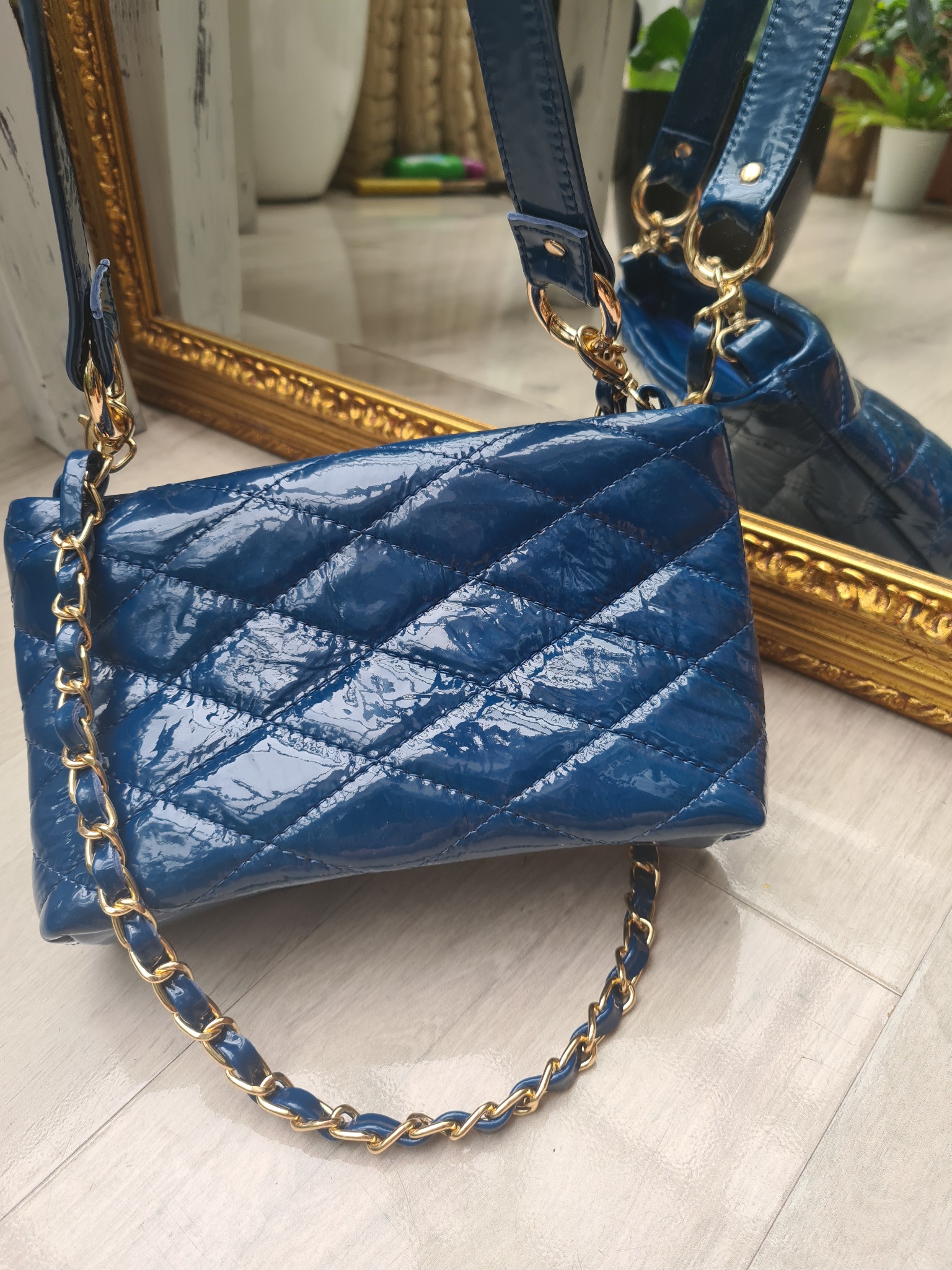 Błyszcząca, pikowana, granatowa torebka damska z łańcuszkiem na tle lustra w złotej ramie.
