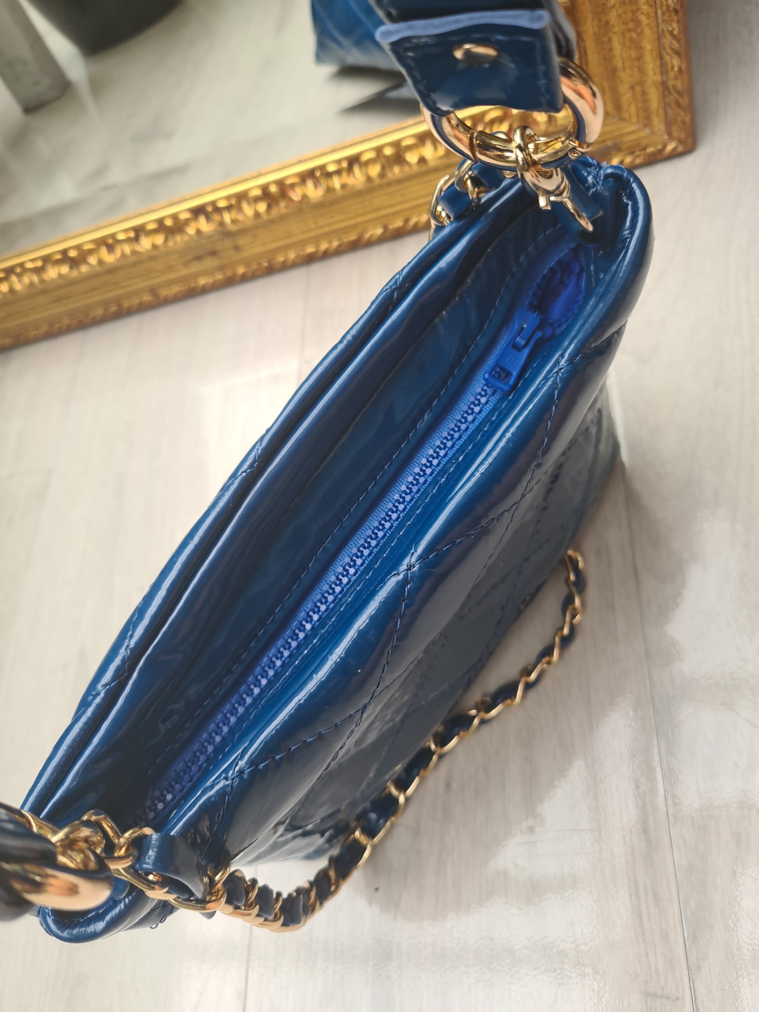 Błyszcząca, pikowana, granatowa torebka z złotym łańcuszkiem, widoczny niebieski zamek, na tle fragmentu lustra w złotej ramie.