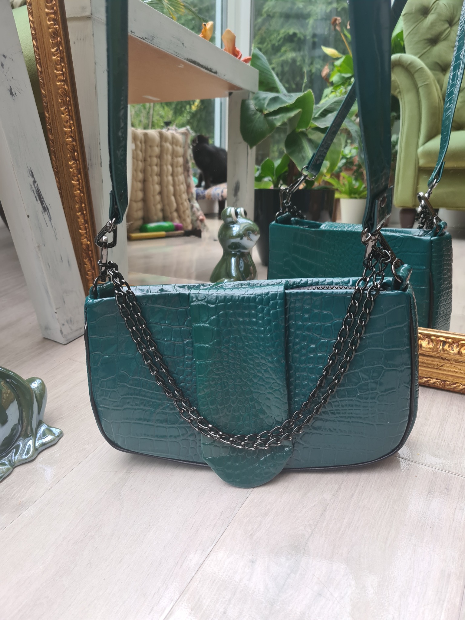 Zielona torebka z imitacji skóry krokodyla, z łańcuszkiem, na jasnej podłodze, w tle rośliny i dekoracje.