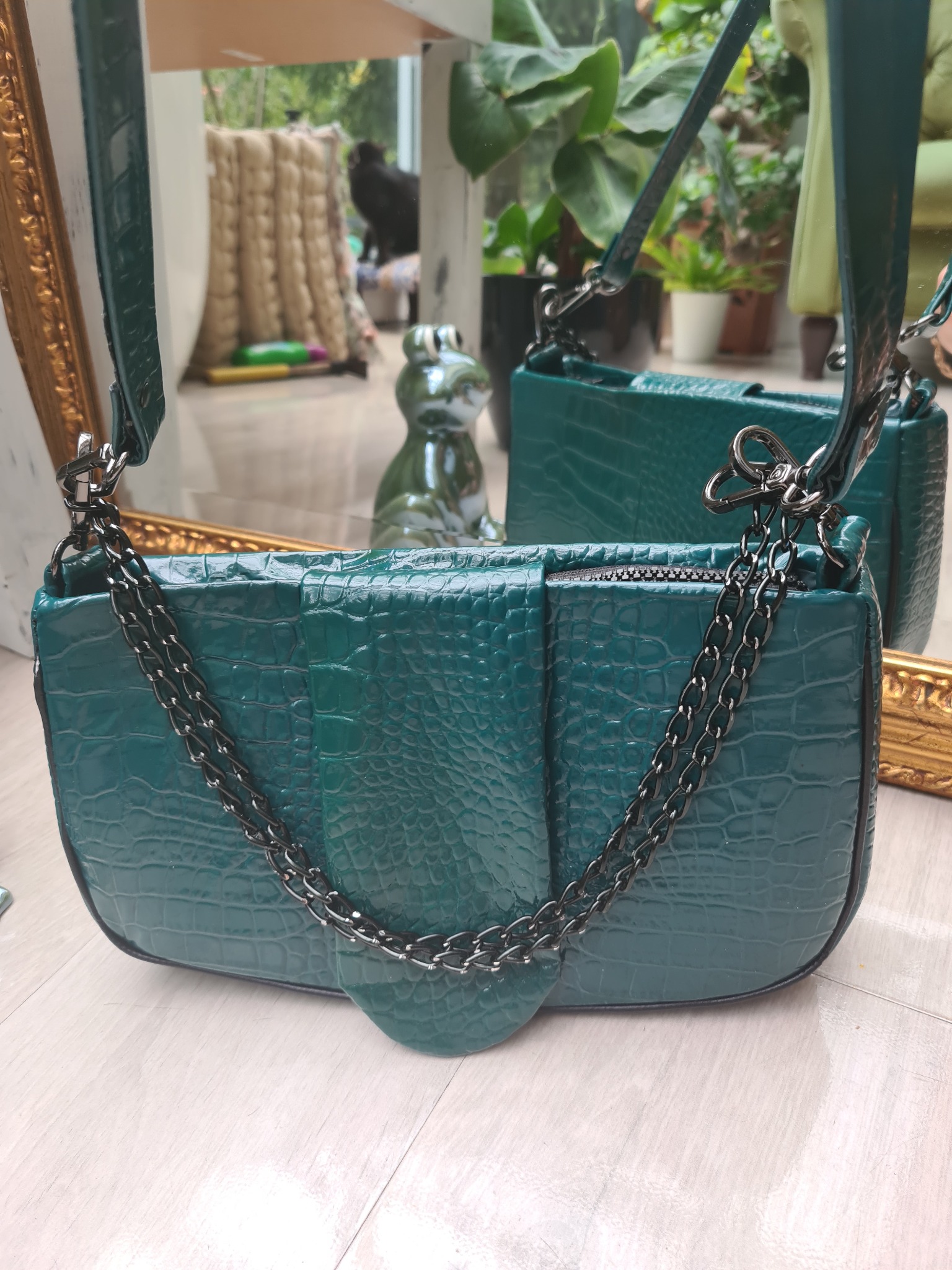 Zielona torebka z imitacji skóry krokodyla, łańcuszek w kolorze grafitowym, odbicie w lustrze z ozdobną ramą, w tle figurka żaby i rośliny.