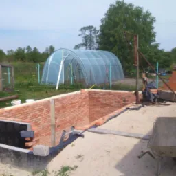Budowa piwnicy z cegły w Stanisławowie Drugim, widoczne fundamenty, izolacja przeciwwilgociowa oraz częściowo wymurowane ściany, w tle szklarnia.