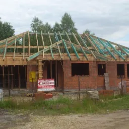 Niedokończony budynek jednorodzinny z czerwonej cegły, z drewnianą konstrukcją dachu w trakcie budowy, widoczne ściany bez okien, teren budowy z materiałami i ogrodzeniem.