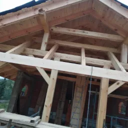 Konstrukcja drewniana dachu w trakcie budowy, widoczne krokwie, łaty i murłat, ceglane ściany z okrągłymi oknami, rusztowanie robocze w tle zieleń.