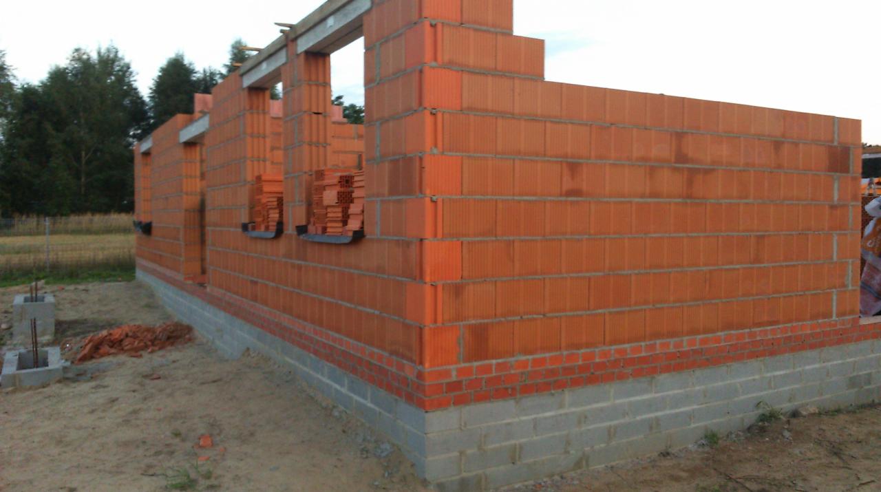 Wznoszone ściany zewnętrzne domu z czerwonej ceramiki poryzowanej, widoczne otwory okienne z przygotowanymi nadprożami, fundament częściowo obłożony cegłą klinkierową, teren budowy z piaskiem.