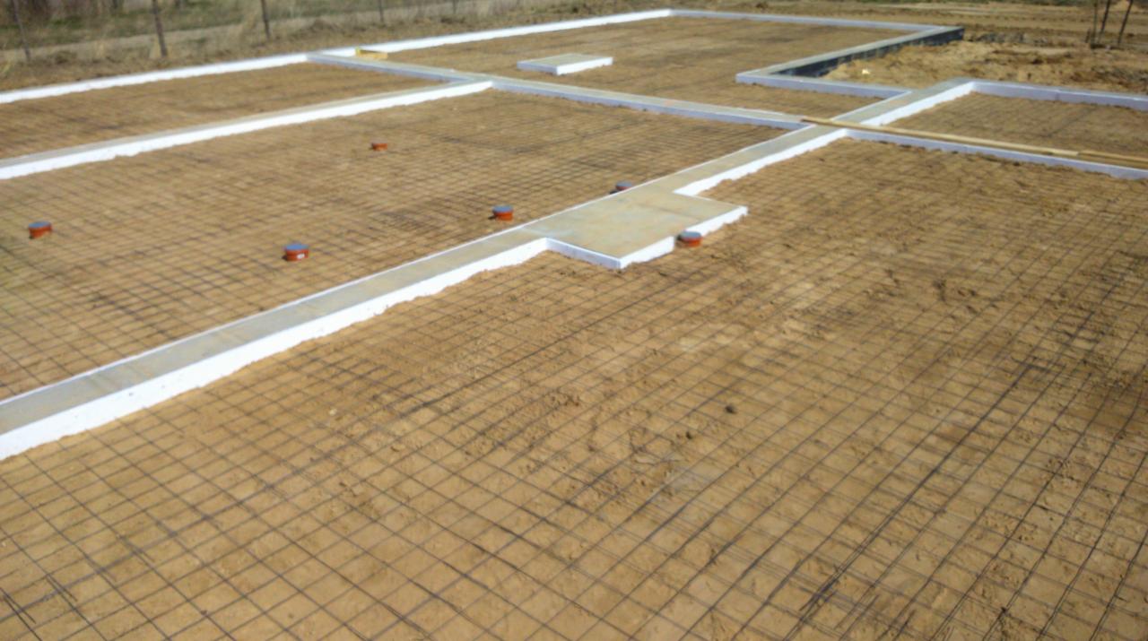 Przygotowany teren pod budowę z ułożoną siatką zbrojeniową i obrysem szalunków fundamentowych, widoczne wystające pionowe rury.