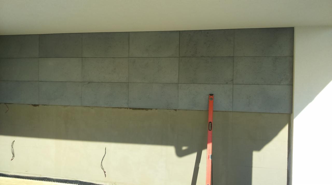 Fragment ściany budynku w trakcie wykończenia z szarymi płytami elewacyjnymi, widoczny poziomica, cień i fragment niezakończonej powierzchni poniżej.