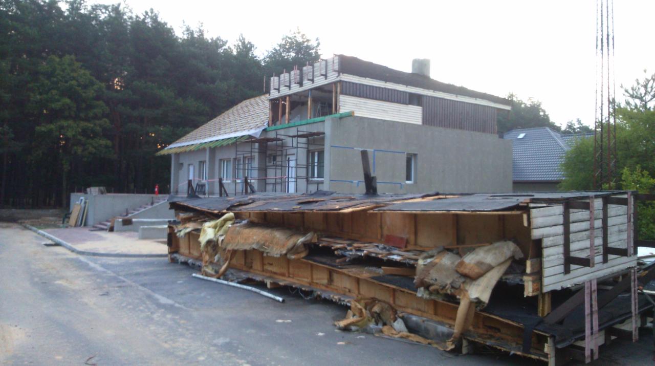 Wieloetapowa budowa domu z elementami murowanymi i drewnianymi, widoczne rusztowania, częściowo położony dach oraz kontener ze starymi elementami konstrukcyjnymi na pierwszym planie.