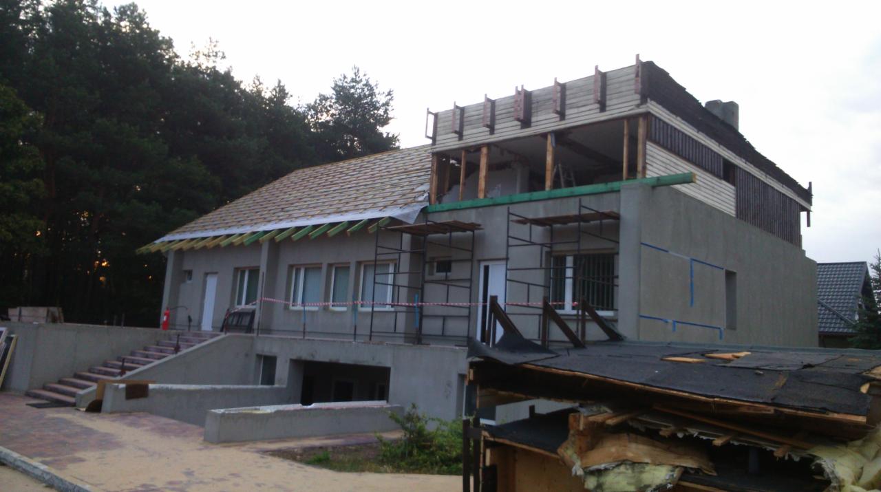 Rozbudowa domu murowanego z częściowo zdjętym dachem, widoczne rusztowania i elementy konstrukcyjne, w tle las.