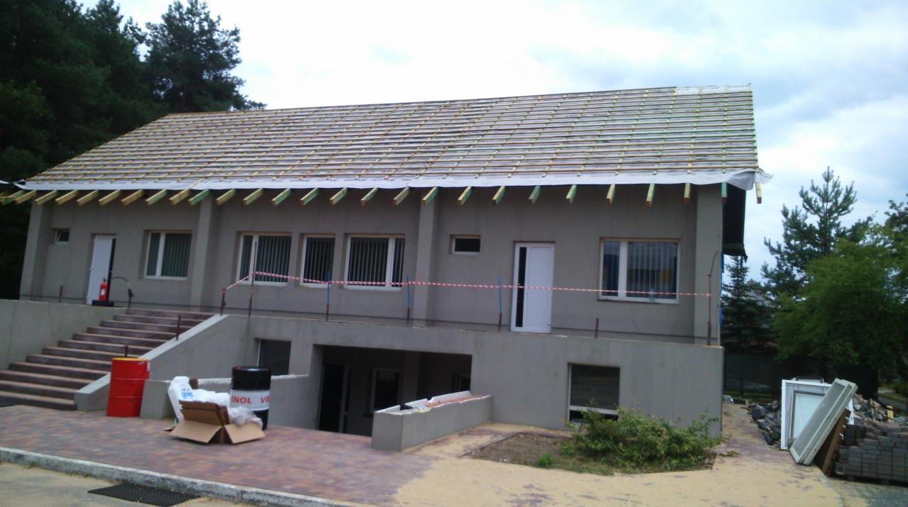 Dwukondygnacyjny budynek w trakcie budowy dachu, widoczne krokwie i membrana dachowa, z boku budynku leżą nowe okna i kostka brukowa, a na schodach stoi czerwona beczka.