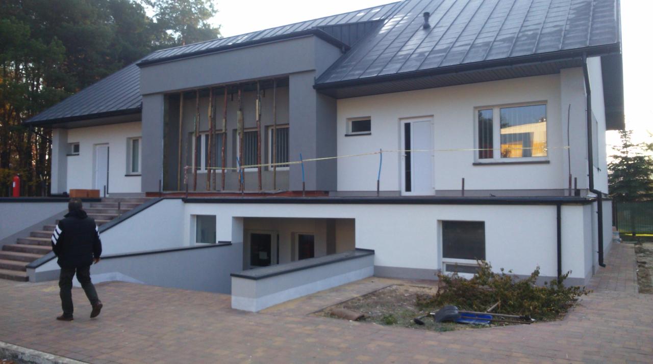 Dwukondygnacyjny budynek z płaskim dachem w trakcie prac wykończeniowych; widoczne rusztowania na elewacji, częściowo zagospodarowany teren wokół budynku i osoba w oddali.