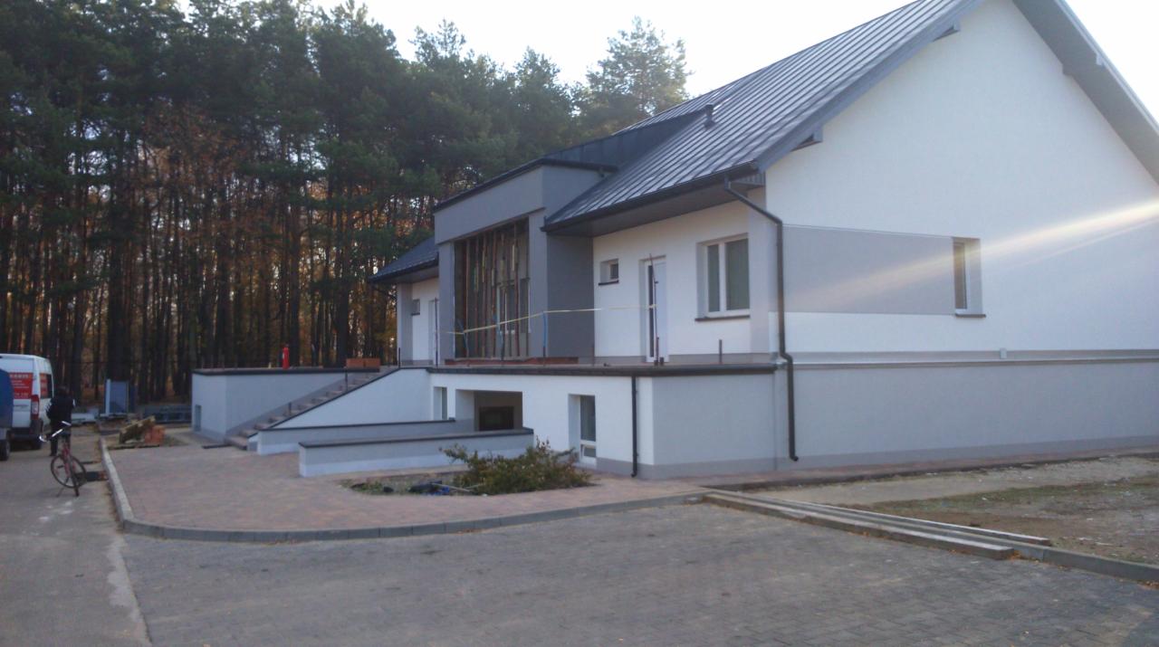 Dwukondygnacyjny budynek z szarą blachodachówką, częściowo wykończony, z widocznymi elementami rusztowania na balkonie, otoczony drzewami i brukowanym terenem.