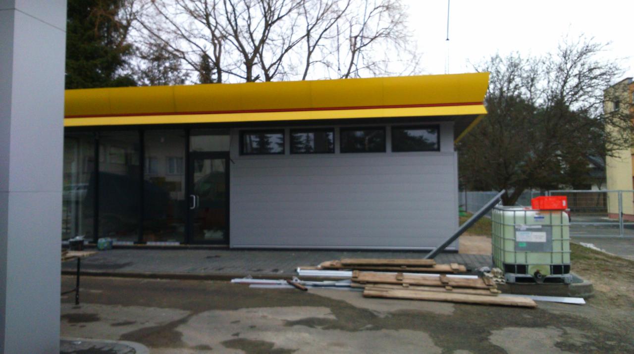 Nowoczesny pawilon handlowy z żółtym zadaszeniem i szarą elewacją, widoczne materiały budowlane i zbiornik na wodę, otoczenie placu budowy.