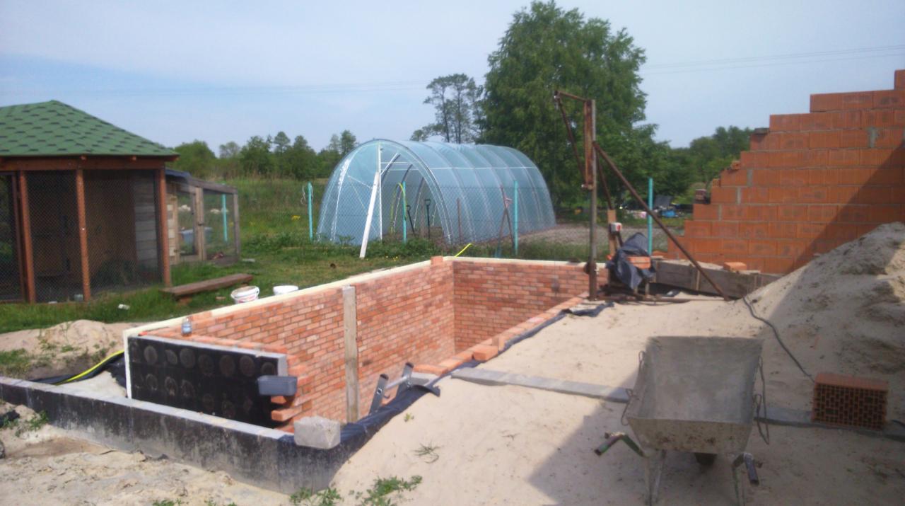 Budowa piwnicy z cegły w Stanisławowie Drugim, widoczne fundamenty, izolacja przeciwwilgociowa oraz częściowo wymurowane ściany, w tle szklarnia.