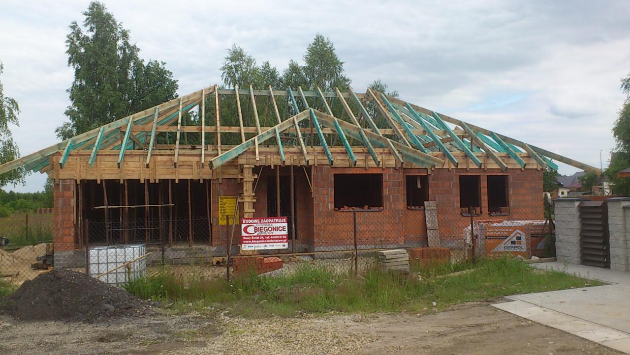 Niedokończony budynek jednorodzinny z czerwonej cegły, z drewnianą konstrukcją dachu w trakcie budowy, widoczne ściany bez okien, teren budowy z materiałami i ogrodzeniem.