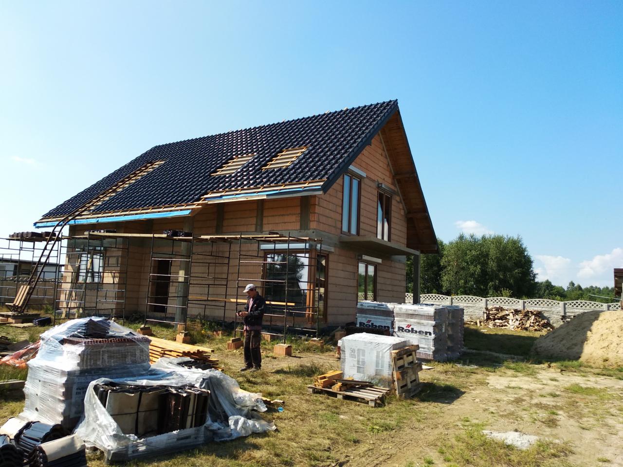 Dom murowany w trakcie budowy z częściowo położonym ciemnym dachem, widoczne rusztowania, palety z dachówką marki Roben oraz pracownik w oddali.