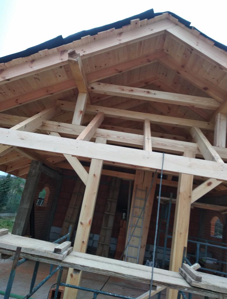 Konstrukcja drewniana dachu w trakcie budowy, widoczne krokwie, łaty i murłat, ceglane ściany z okrągłymi oknami, rusztowanie robocze w tle zieleń.