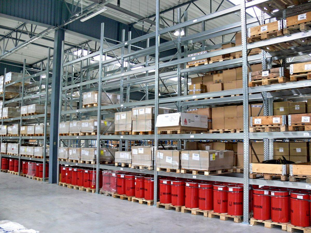 Wysokie, metalowe półki magazynowe wypełnione kartonami, paletami i czerwonymi pojemnikami, widok z perspektywy podłogi magazynu.