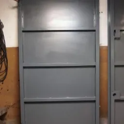 Szara metalowa brama garażowa z drewnianymi uchwytami, oparta o drewniany klocek, widoczna kolejna brama obok.