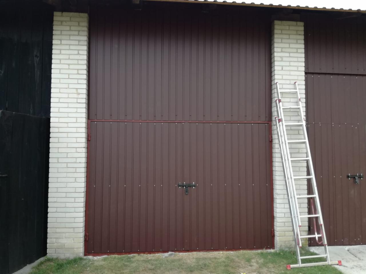 Brązowa brama garażowa z pionowymi przetłoczeniami, obramowana cegłą, z aluminiową drabiną opartą o ścianę, widoczna trawa u podstawy.