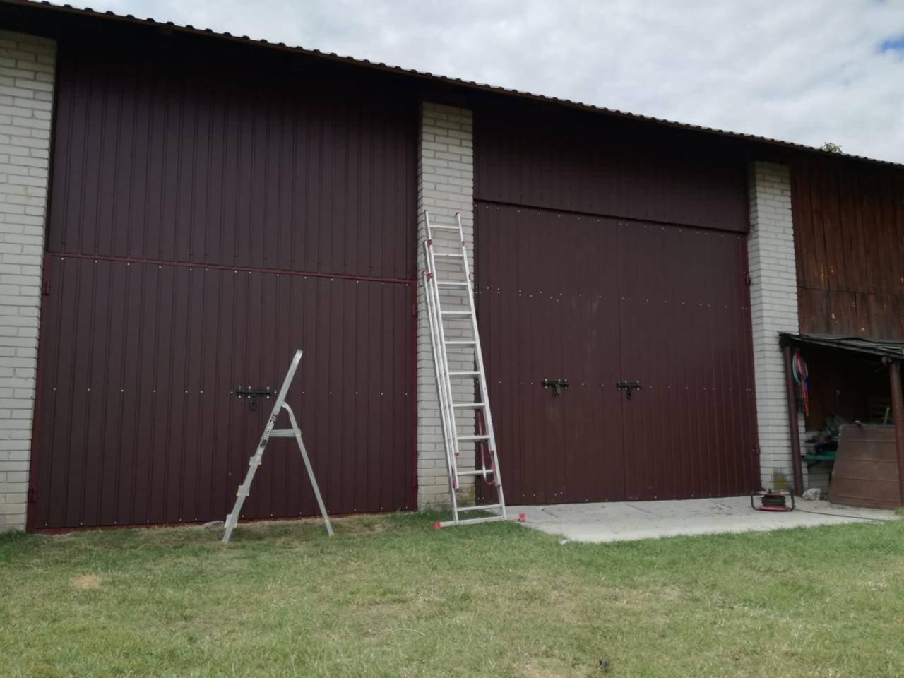 Dwie brązowe, metalowe bramy garażowe z zamkami, osadzone w murze z białej cegły, z dwiema rozstawionymi aluminiowymi drabinami na trawniku przed nimi.