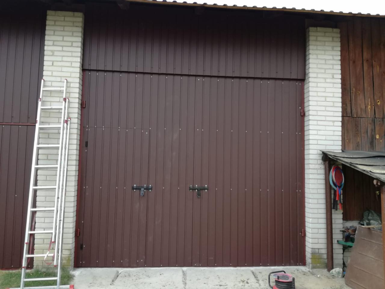 Brązowa, dwuskrzydłowa brama garażowa z pionowymi panelami, zabezpieczona dwoma zamkami, osadzona pomiędzy białymi, ceglanymi filarami, z aluminiową drabiną opartą o jeden z filarów i widocznym...