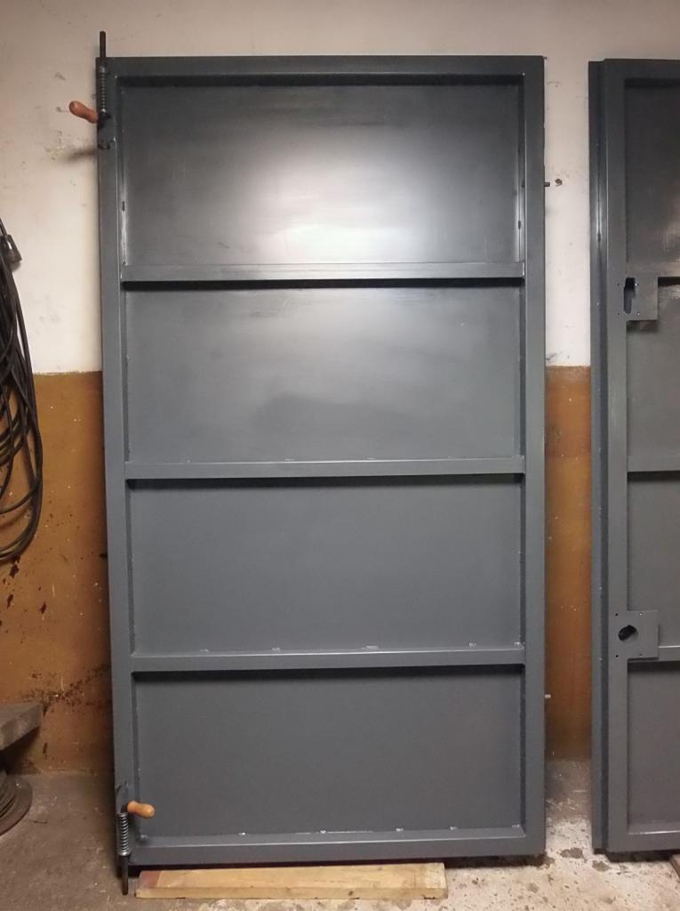 Szara metalowa brama garażowa z drewnianymi uchwytami, oparta o drewniany klocek, widoczna kolejna brama obok.