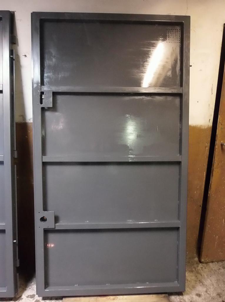 Szara, metalowa rama drzwiowa z widocznymi śladami po spawaniu i niedoskonałościami w malowaniu, stojąca w warsztacie.